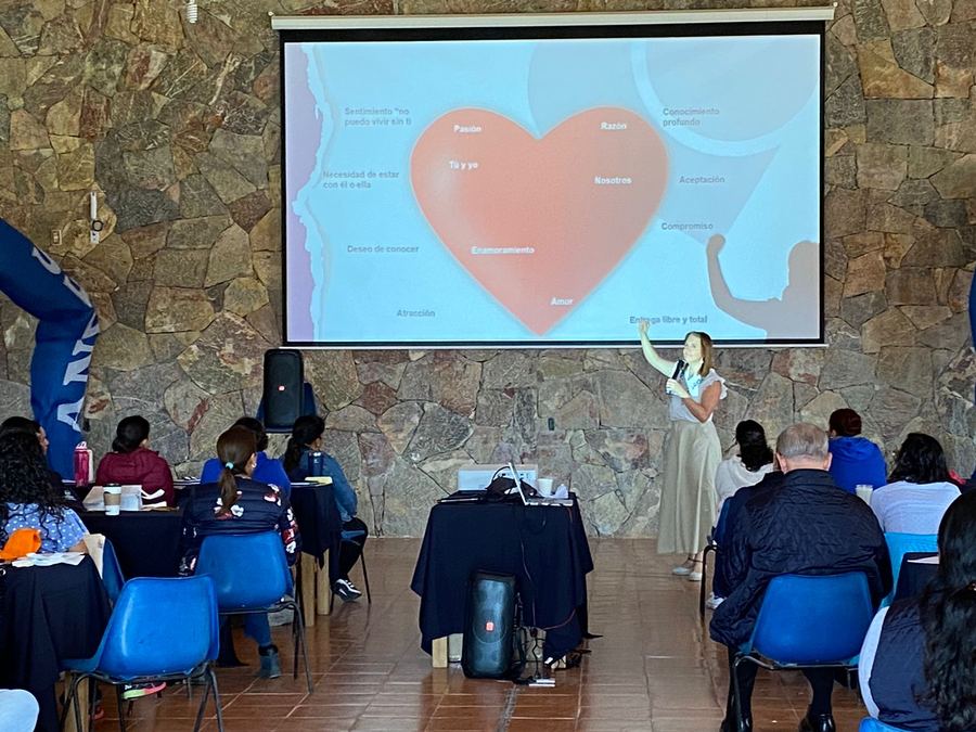 Un salón con muro de piedra lleno de asistentes sentados en mesas mira una pantalla con un corazón mientras Dany explica.