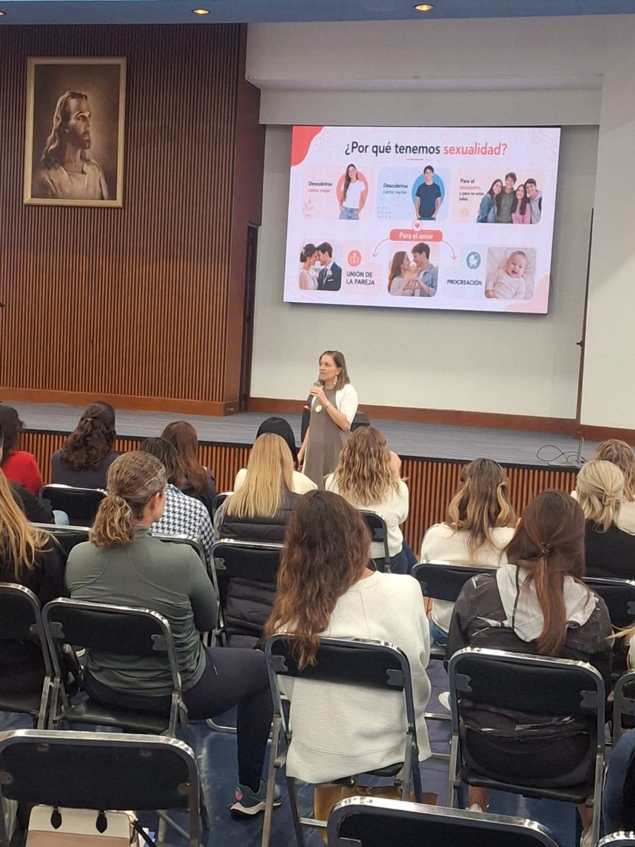 Dany imparte una conferencia a un salón lleno; en la pantalla se lee «¿Por qué tenemos sexualidad?».
