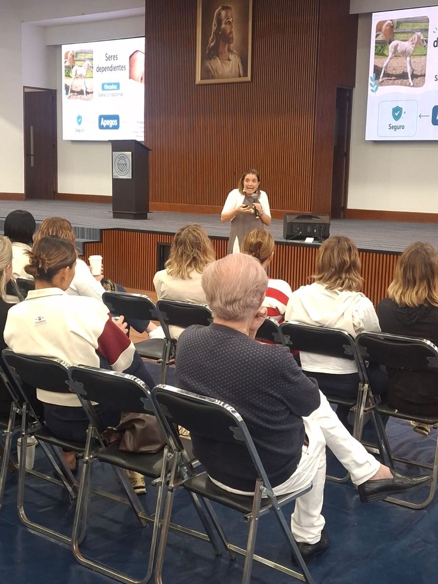 Un auditorio de adultos escucha a Dany, que habla al fondo junto a dos pantallas.